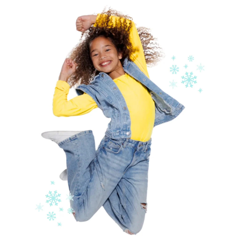 Lachend meisje in geel shirt en spijkerjack maakt een sprong tussen sneeuwvlokken